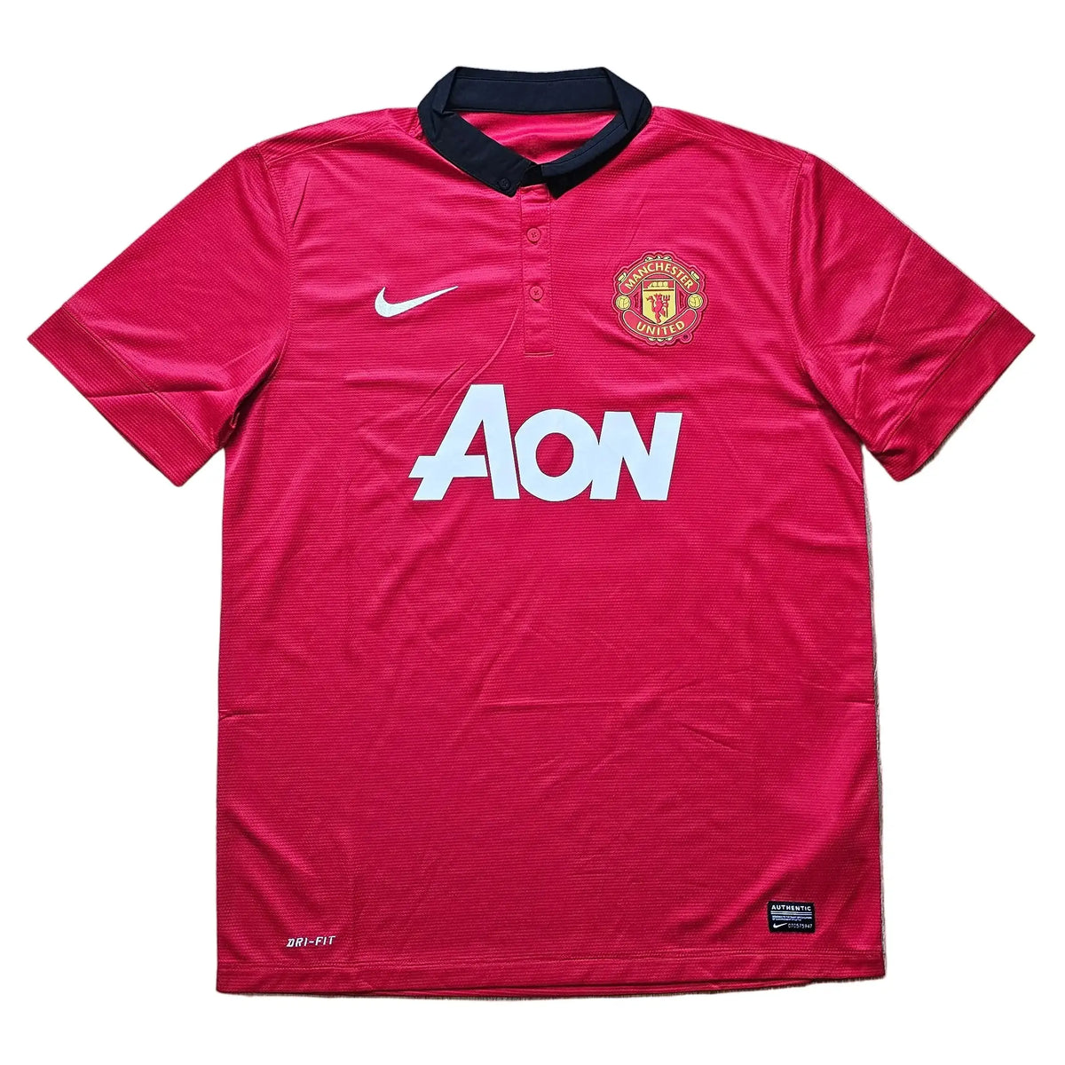 A red Manchester United Nike soccer jersey with a black collar. It features the white AON sponsor logo across the chest, a white Nike swoosh on the right chest, and the Manchester United club crest on the left chest.