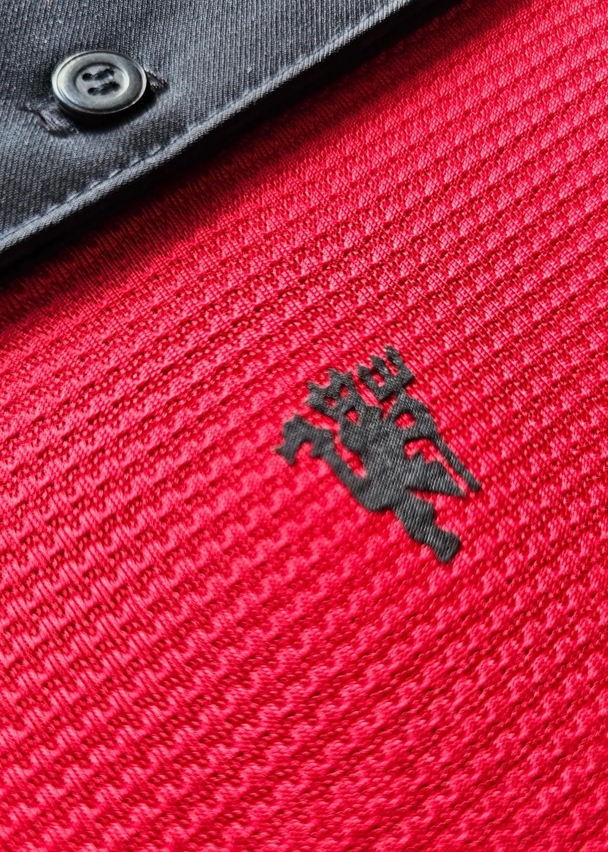 Close-up of a red ribbed knit top with a black embroidered club logo on the chest and a dark gray buttoned collar visible at the top.