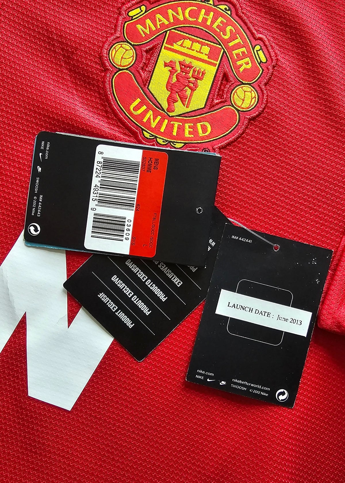 Close-up of a red Manchester United soccer jersey with the club crest visible, along with black hang tags and a white size tag laid on the fabric.